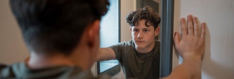 Teenage male looks at himself in a mirror with his hands pressed against the wall.