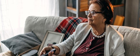 Older woman sits on couch using television remote.
