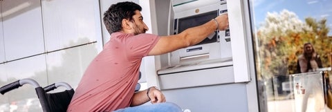 A man in a wheelchair reaches up to an ATM bank machine to insert his banking card.