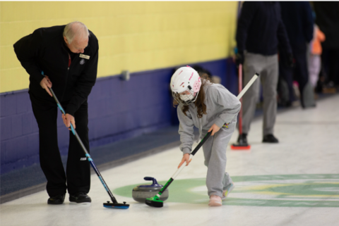 An adult and a child wearing a helmet play curling together on the ice, each holding a broom while guiding a stone