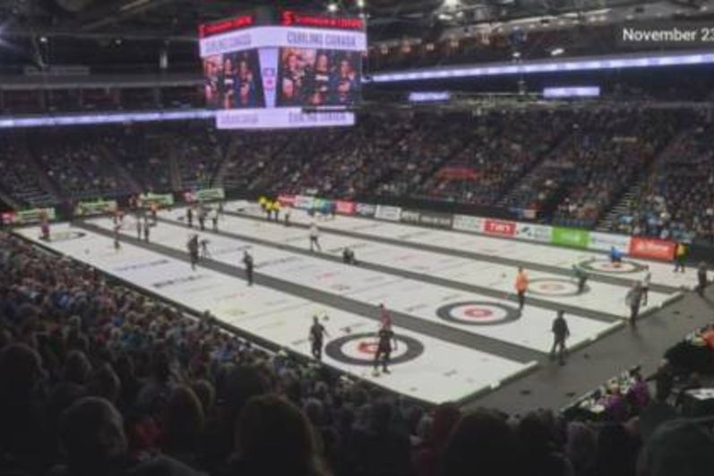 Wide view of a packed arena during a curling competition, showing multiple sheets of ice in use with athletes, officials, and spectators filling the stands, and a large scoreboard hanging above the ice.