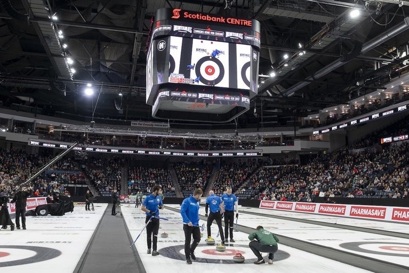 Curling teams compete on the ice inside a sold-out arena, with players delivering stones while teammates sweep and a large overhead scoreboard above the sheets.