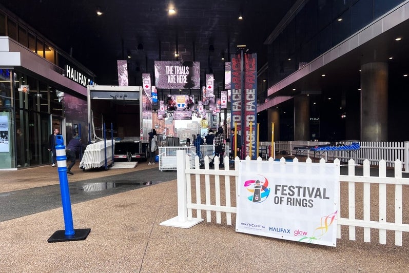 Outdoor entrance to the Festival of Rings event in Halifax, with white fencing, event signage, and banners reading the trials are here inside a covered walkway as staff set up the venue.