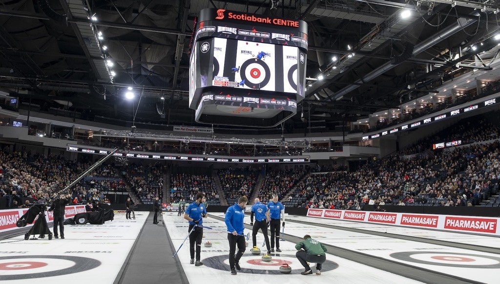Curling teams compete on the ice inside a sold-out arena, with players delivering stones while teammates sweep and a large overhead scoreboard above the sheets.