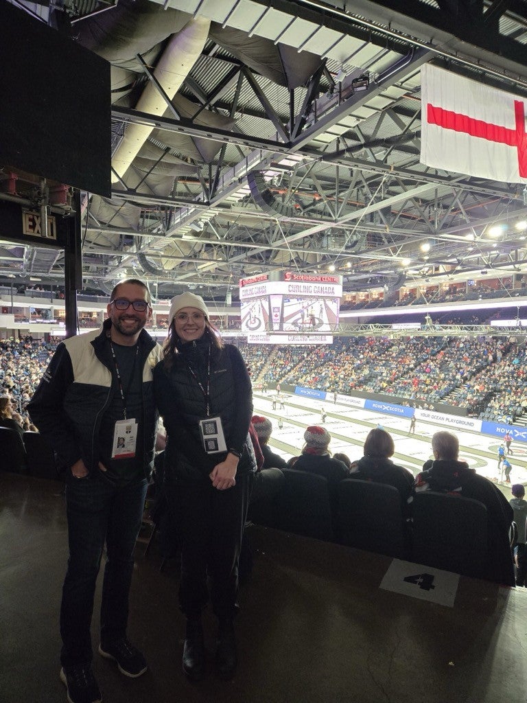 Dr. Simon Barrick and Dr. Kristi Allain wearing event credentials stand in front of a packed arena with multiple curling sheets visible behind them. 