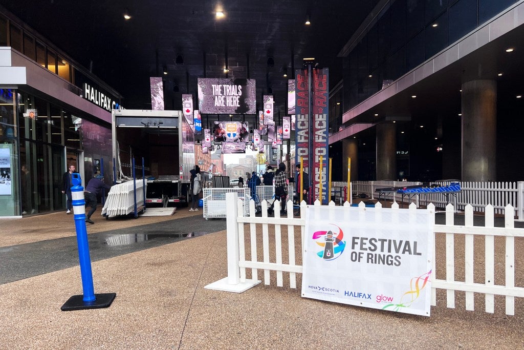 Outdoor entrance to the Festival of Rings event in Halifax, with white fencing, event signage, and banners reading the trials are here inside a covered walkway as staff set up the venue.