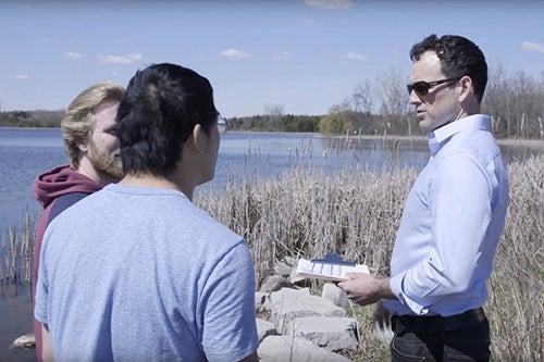 Professor Bryan Grimwood talks to two students at shore of a lake.