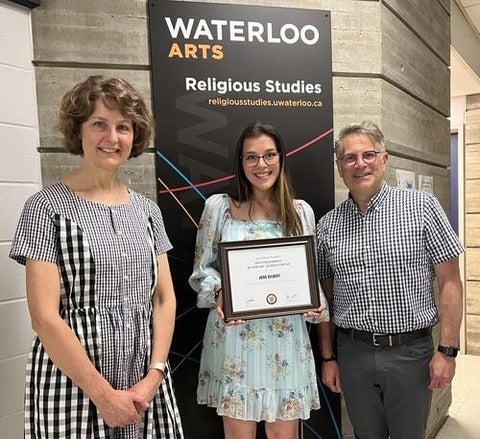 Jess Gilbert holds up her award infront of the Religious Studies sign, posing with two faculty members