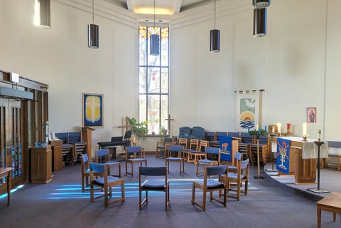 A chapel interior with wooden chairs, candles, banners, and a tall stained-glass window.