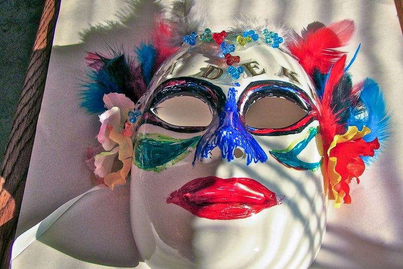 A decorated white mask with colourful paint, feathers, flowers, and beads.