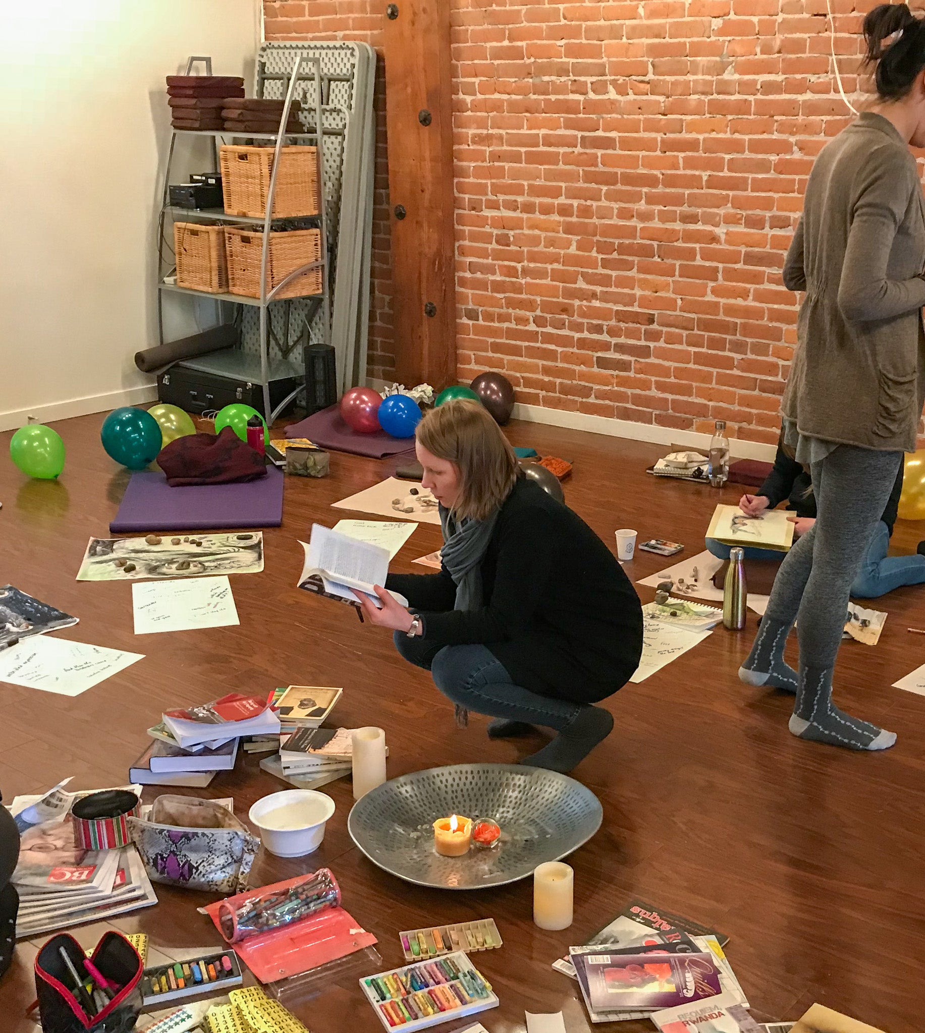 People sitting on the floor with art supplies, papers, books, candles, and balloons in a workshop space.