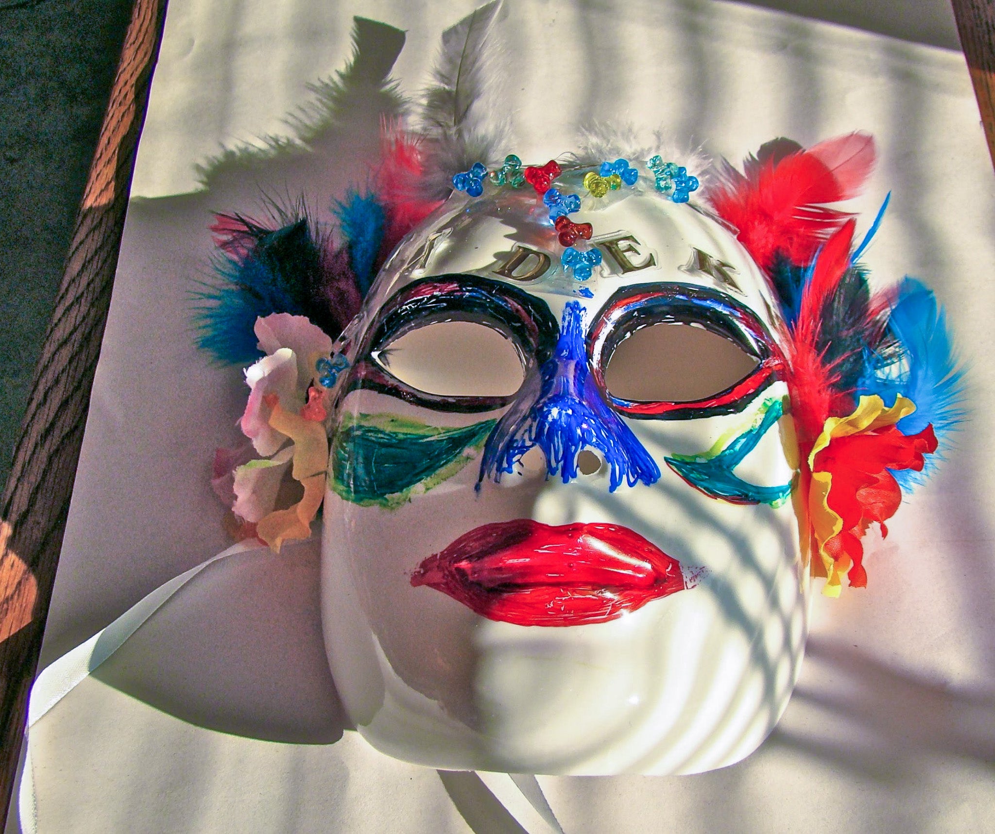 A decorated white mask with colourful paint, feathers, flowers, and beads.