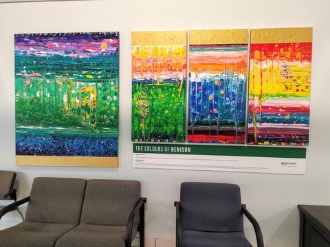 Four paintings handing on a wall. The one on the left is large standalone painting and has some dark blues, purples and greens with a smattering of lighter colour. There is a triptych shown on the right. Each painting has a gold bar across, and has a rainbow of colours to represent Renison employees. 