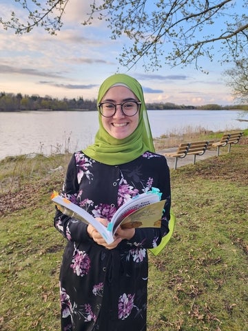 Aurra Startup holding a book open in front of the river. She is wearing a green headscarf and glasses, and is smiling at the camera. 