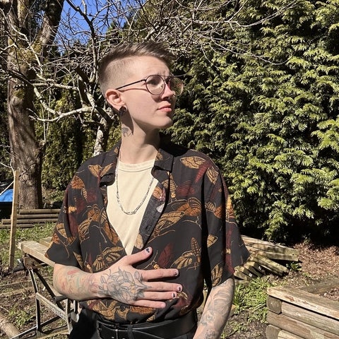 Alex standing in front of a tree. They are wearing a short sleeve button up shirt and glasses. 