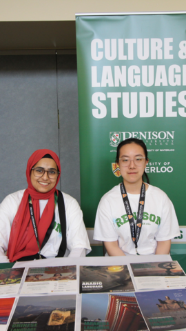 Two people sitting at a table at a recruitment event with a Culture and Language Studies banner behind them.