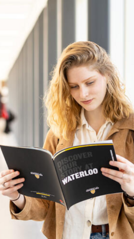 Person standing near a wall of windows reading a uWaterloo Viewbook.