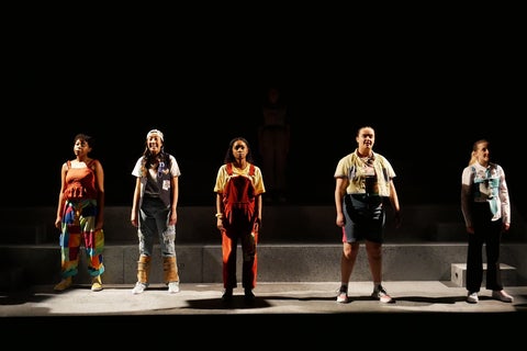 Stage lights illuminate an ensemble of actresses of diverse ethnicity standing in a line facing the audience. The actresses all depict a single character in the play ‘Red Bike’ by Caridad Svich. And yet their expressions are as varied and as unique as their colourful theatrical attire. 