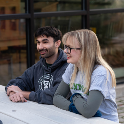 Two students sitting together at a picnic table. They are leaning on the table, having a conversation. 