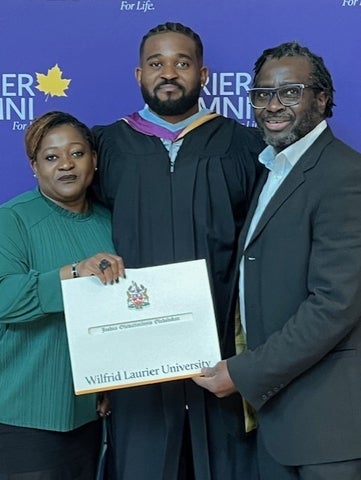Three people standing in front of a Laurier backdrop. The middle person is wearing graduation robes and is holding a degree. 