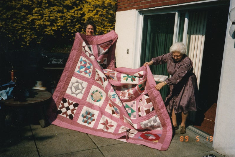Two women hold up a quilt made up of several squares, each with a different pattern. It is a dusty rose colour with some blues and yellows and reds. 