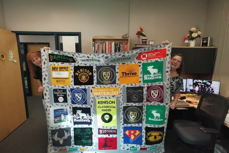 Brenda's most recent quilt, which has been created with the fronts of t-shirts from Renison through the years. The quilt is being held up by Brenda and a colleague. 