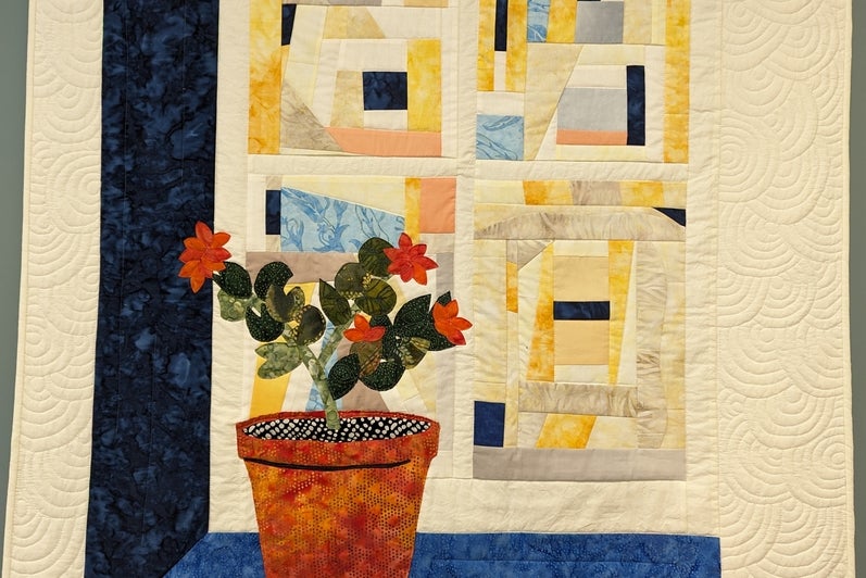 Quilt with a beige background and potted flowers in the foreground. It appears 3-dimensional because of the blue wall and counter beside and beneath the flowers. The background has squares that are abstract patterns. 