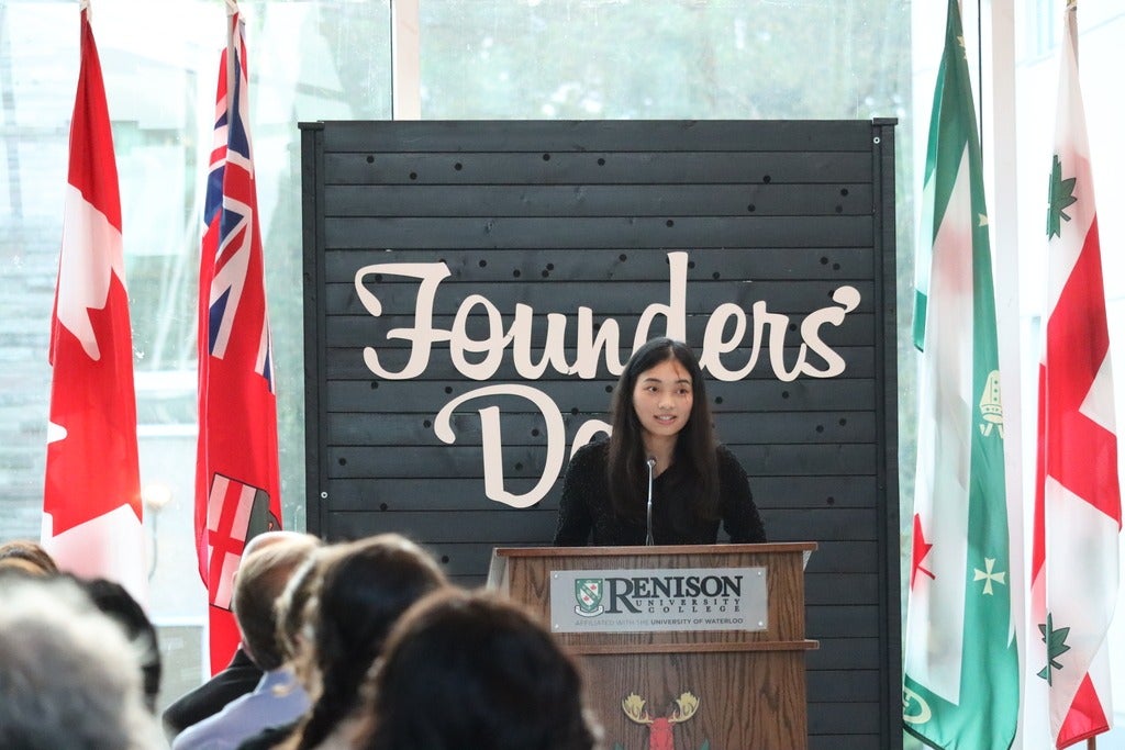 Young woman stands at the Renison podium speaking. She is in front of a large black backdrop that has the words "Founders' Day" on it in white. 