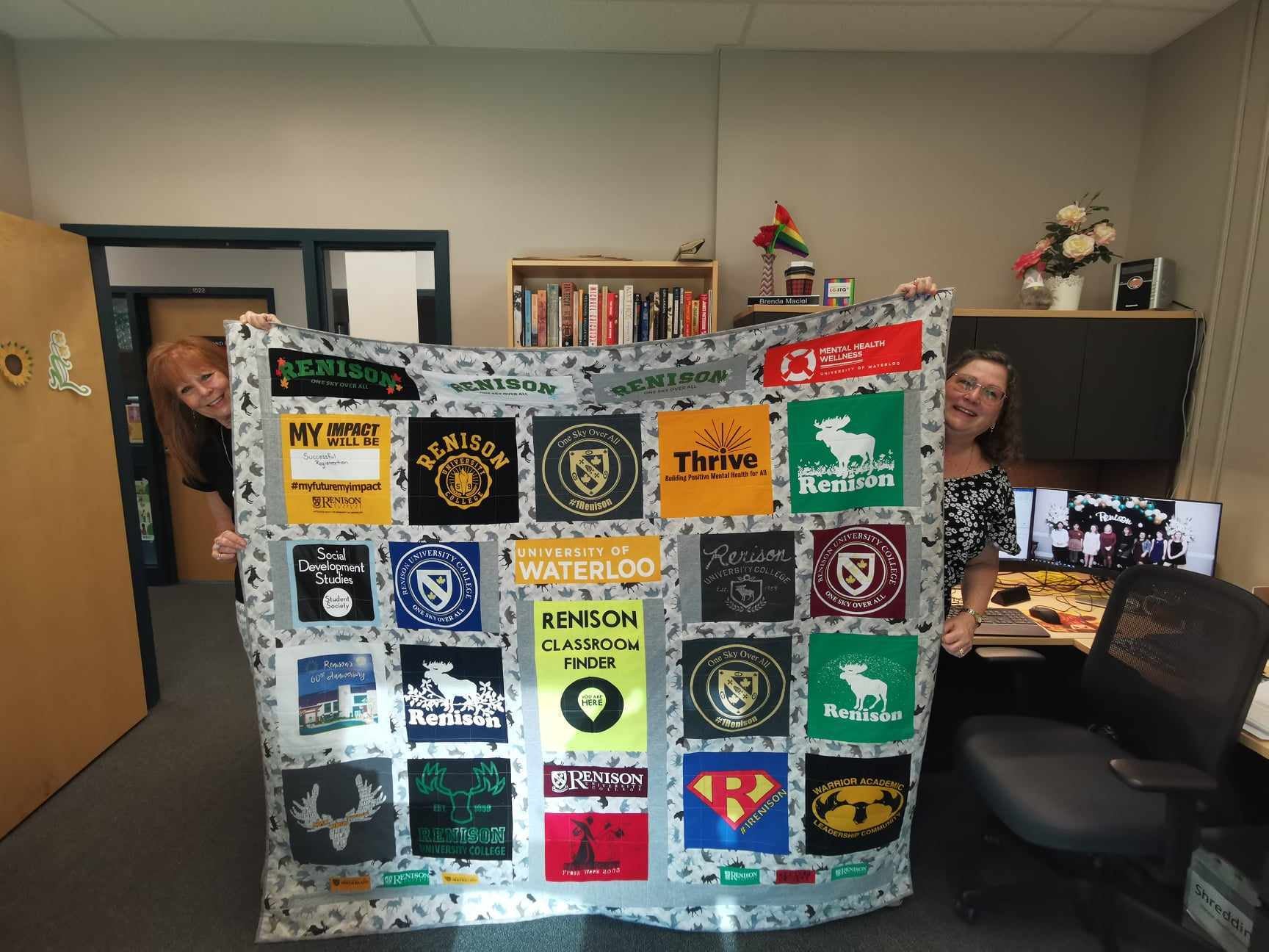 Brenda's most recent quilt, which has been created with the fronts of t-shirts from Renison through the years. The quilt is being held up by Brenda and a colleague. 