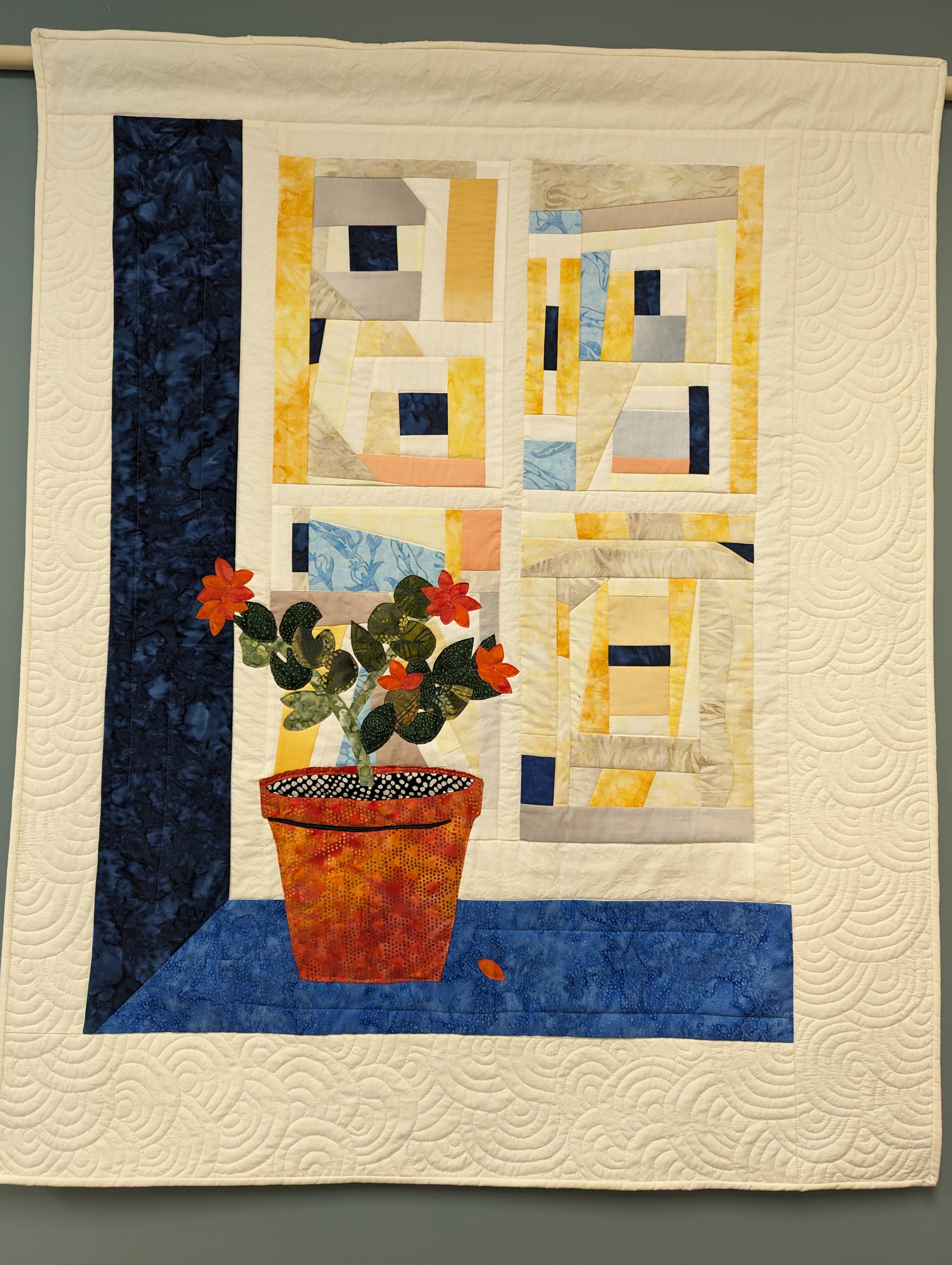 Quilt with a beige background and potted flowers in the foreground. It appears 3-dimensional because of the blue wall and counter beside and beneath the flowers. The background has squares that are abstract patterns. 