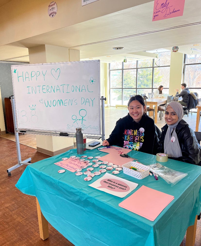 Two students sitting at a table beside a board that reads Happy International Women's Day