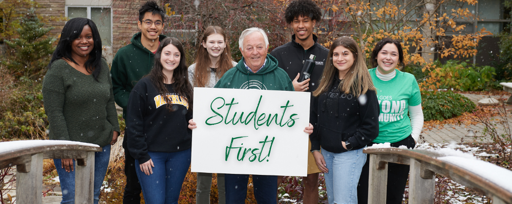 Chancellor Manfred Conrad standing outside with a group of students. Manfred is holding a sign that reads 'Students First'.