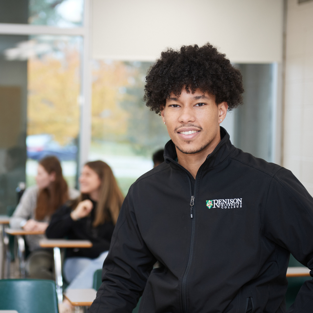 One student wearing a Renison sweatshirt stands smiling at the front of the frame. Students can be seen in the background sitting at desks.