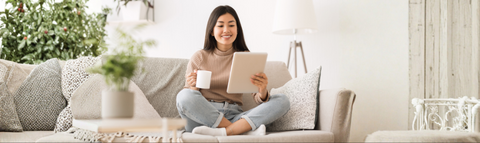Young Asian woman sitting on a couch reading a tablet and holding a white mug