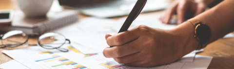 A table with papers, eye glasses, and a white cup and a person holding a pen