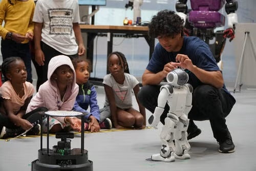 A NAO robot and Turtlebot 4 robot with an Engineering Outreach Leader on the floor in front of a group of kids