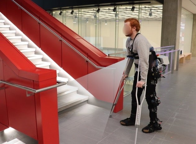 Bearded man using a lower-body exoskeleton and crutches with a chest-mounted camera near stairs