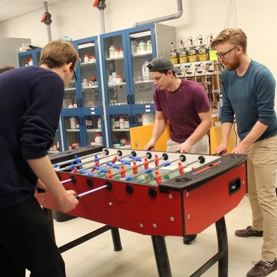 Three students relaxing and playing foosball in the non-active part of the lab
