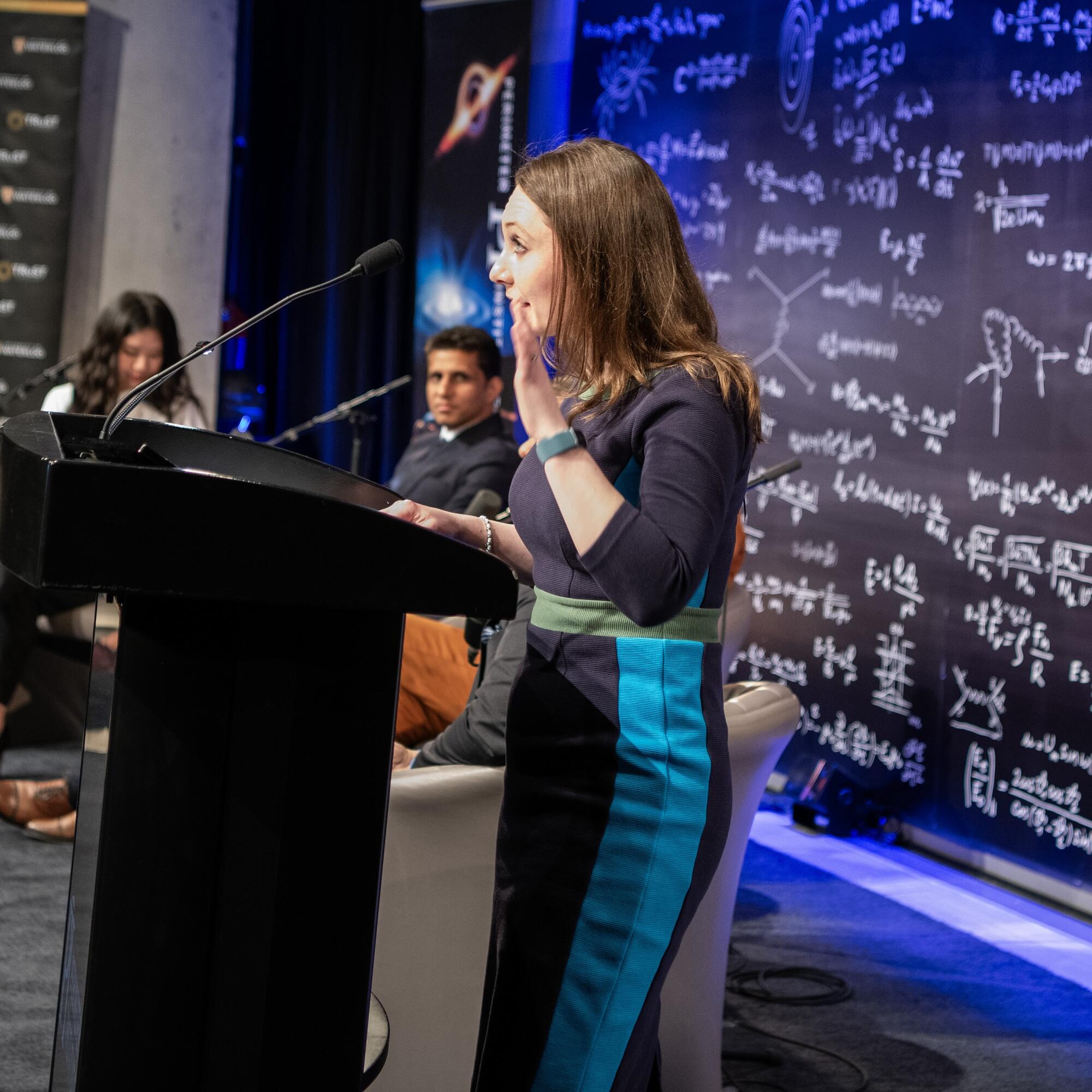 Ashley Rose Mehlenbacher standing at a podium at the Perimeter Institute for Theoretical Physics introducing an event.