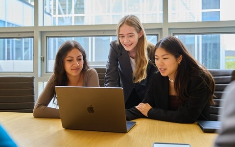 Three students at a boardroom table, working together on a laptop.