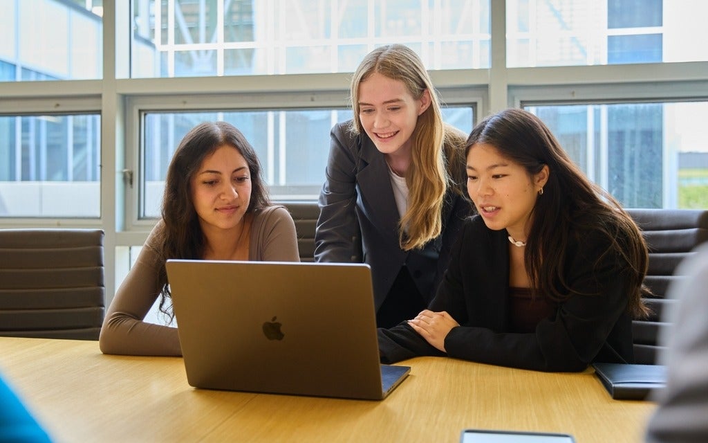 Three students at a boardroom table, working together on a laptop.
