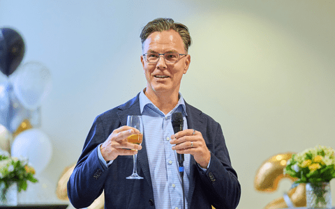 Dr. Blake Phillips gives a toast with a champagne flute and microphone in his hands. There are balloons and flowers in the background.