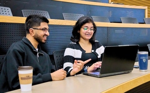 Two students sitting in a classroom and working together on a laptop.
