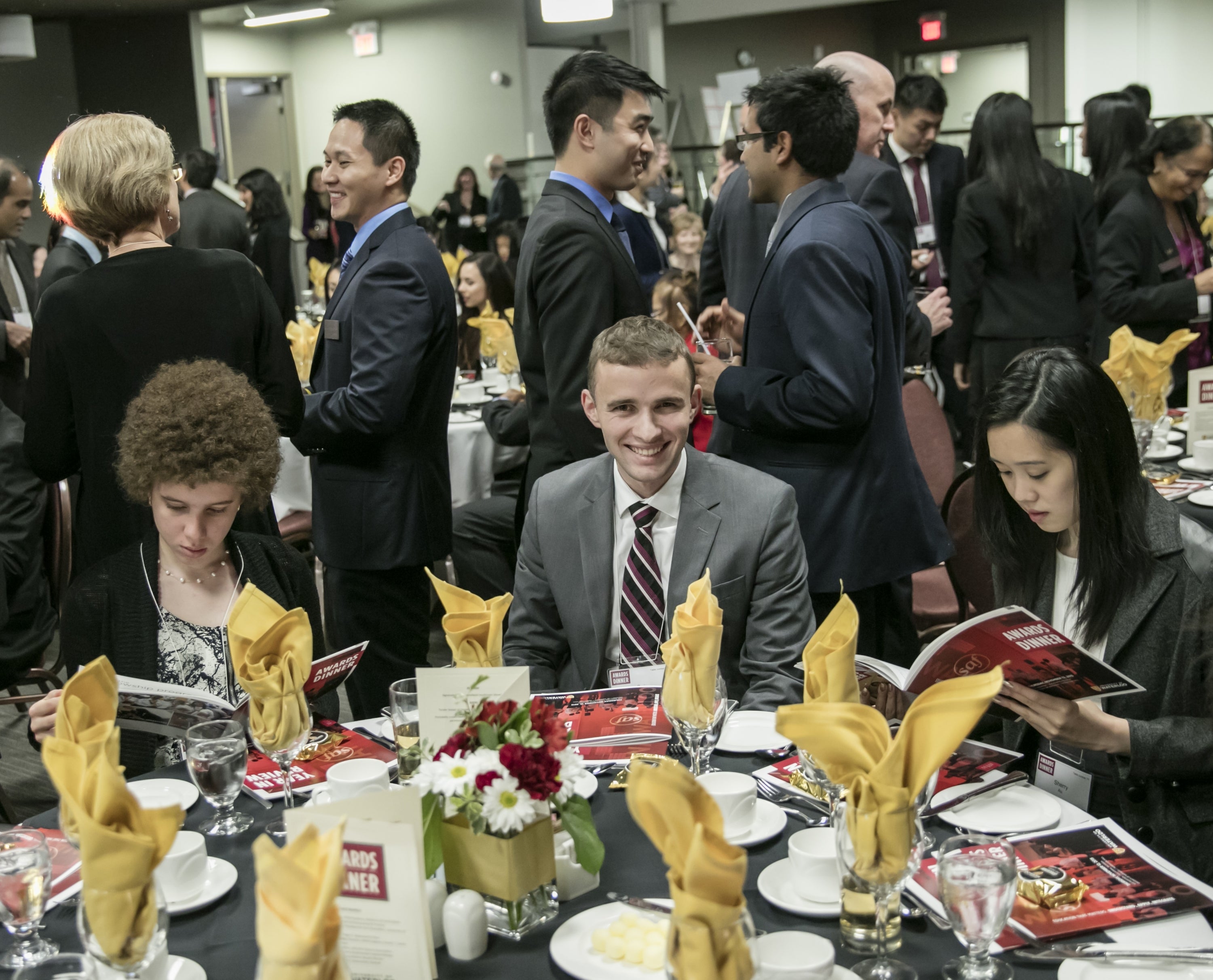 Students sitting at a table, with others in the background mingling