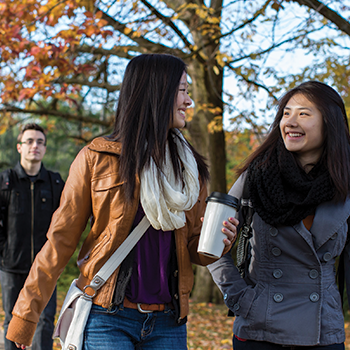 two female students in the foreground and 1 male student in the background walking on a fall day directing to information for future students 