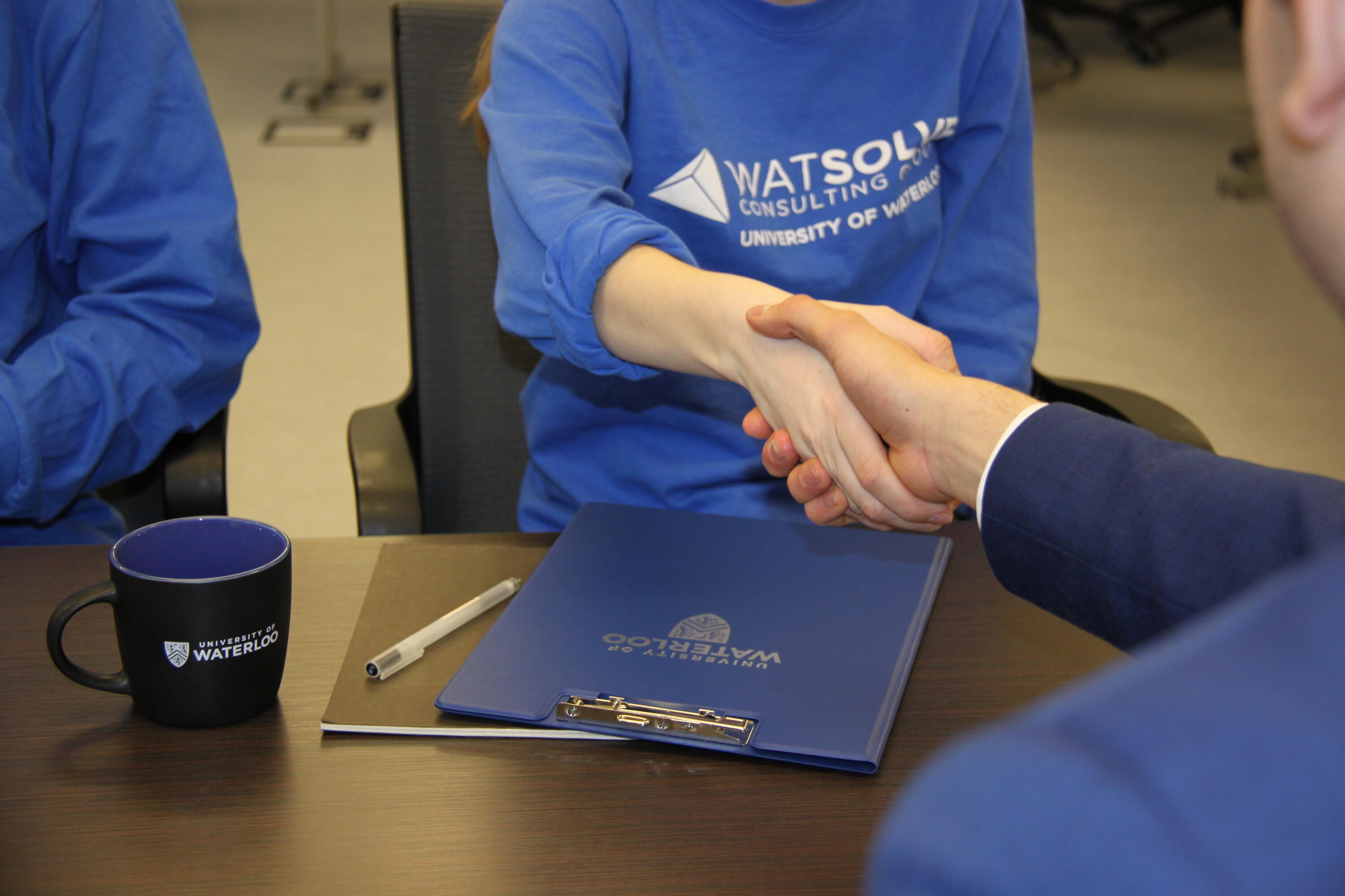 Handshake with blue Waterloo branded mug and notebook on the table.