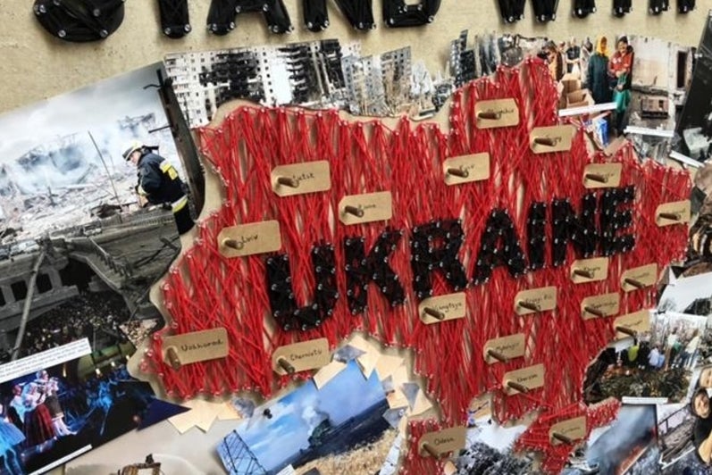 A collage of photos of Ukraine on a wall, with the words “Stand with Ukraine” in black cutout letters attached to the wall and collage.