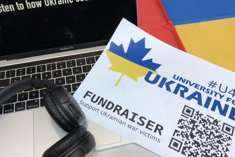 A laptop, headphones, and piece of paper with a QR link to a fundraiser for Ukraine; laptop shows the words “Listen to how Ukraine sounds now”.