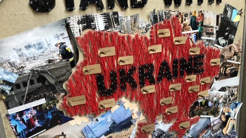 A collage of photos of Ukraine on a wall, with the words “Stand with Ukraine” in black cutout letters attached to the wall and collage.