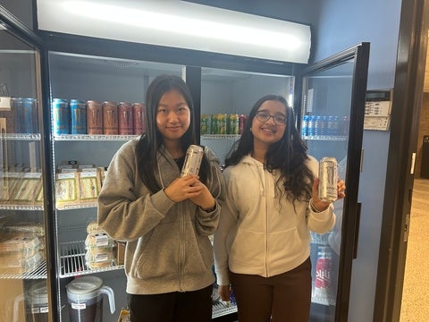 Two people holding energy drinks and posing in front of a fridge